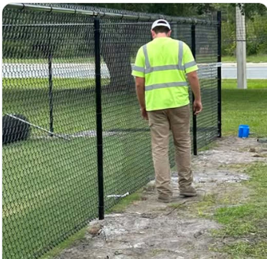 Lowman crew member in a high-visibility vest installing black chain link at a ball field